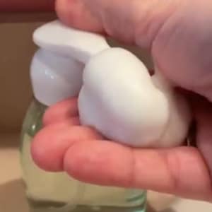 May include: A clear glass bottle with a white pump dispenser. The bottle is filled with a clear liquid and a white foam is being dispensed from the pump.