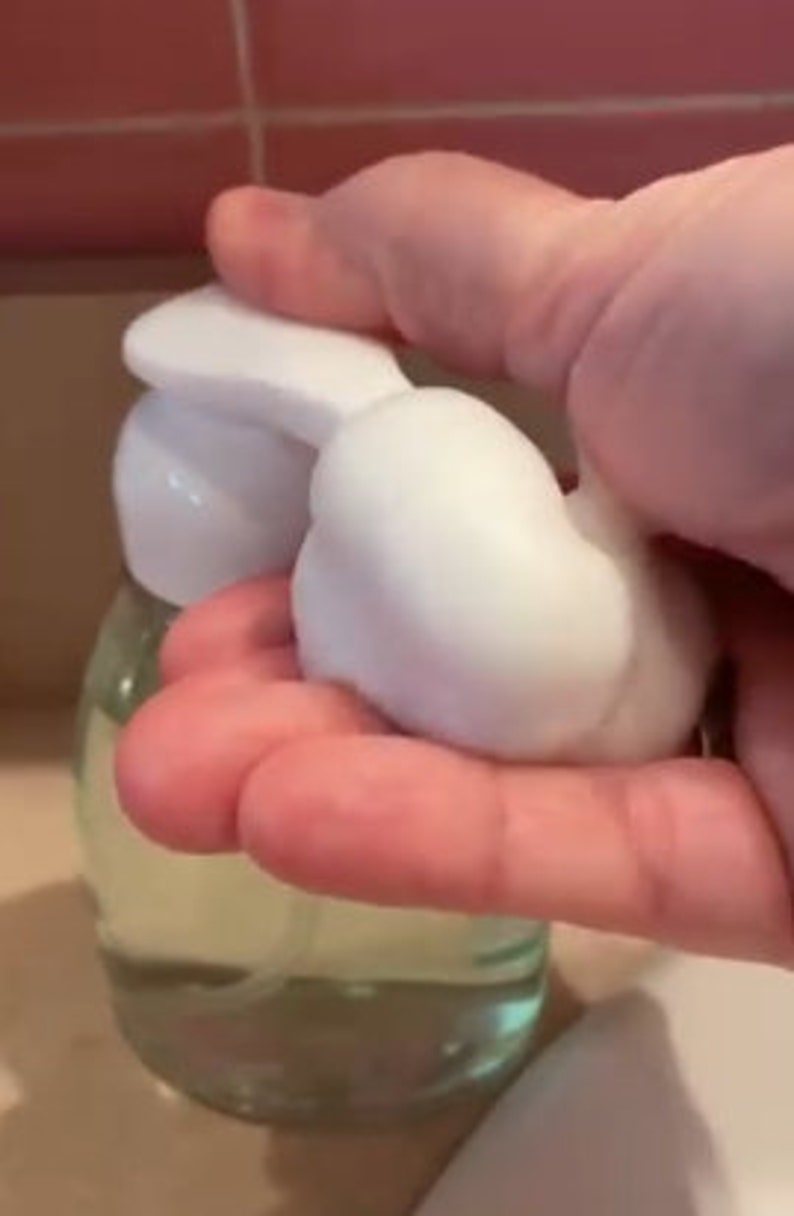 May include: A clear glass bottle with a white pump dispenser. The bottle is filled with a clear liquid and a white foam is being dispensed from the pump.