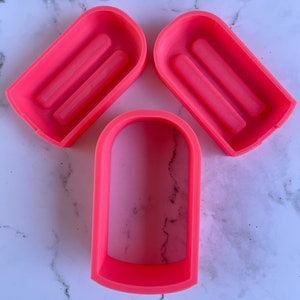 May include: Three pink plastic molds for making soap or candles. The molds are shaped like a rectangle with rounded edges. Two of the molds have a raised design on the inside.