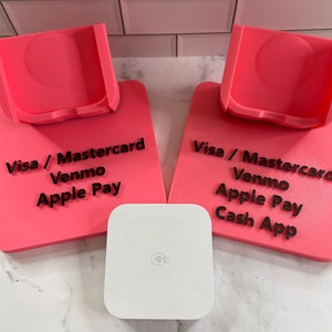 May include: Two pink payment stands with black text that reads "Visa / Mastercard, Venmo, Apple Pay" and "Visa / Mastercard, Venmo, Apple Pay, Cash App". A white square payment device is in front of the stands.