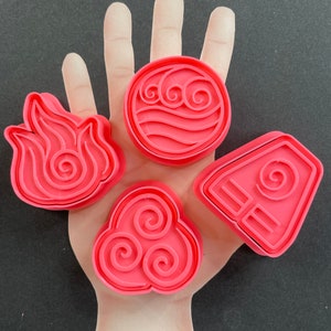 May include: Four pink plastic cookie cutters shaped like the elements from Avatar: The Last Airbender. The cutters are for water, earth, fire, and air.