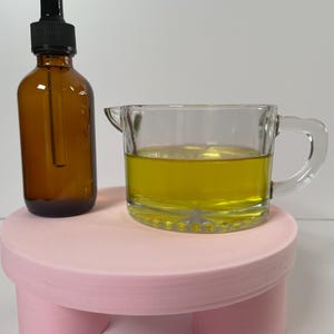 May include: A small, clear glass pitcher with a handle, filled with a yellow liquid, and a brown glass bottle with a dropper, both sitting on a pink, three-legged stand.