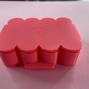 May include: Pink silicone mold with a wavy edge and a small chef logo in the center. The mold is designed for making multiple small cakes or pastries.