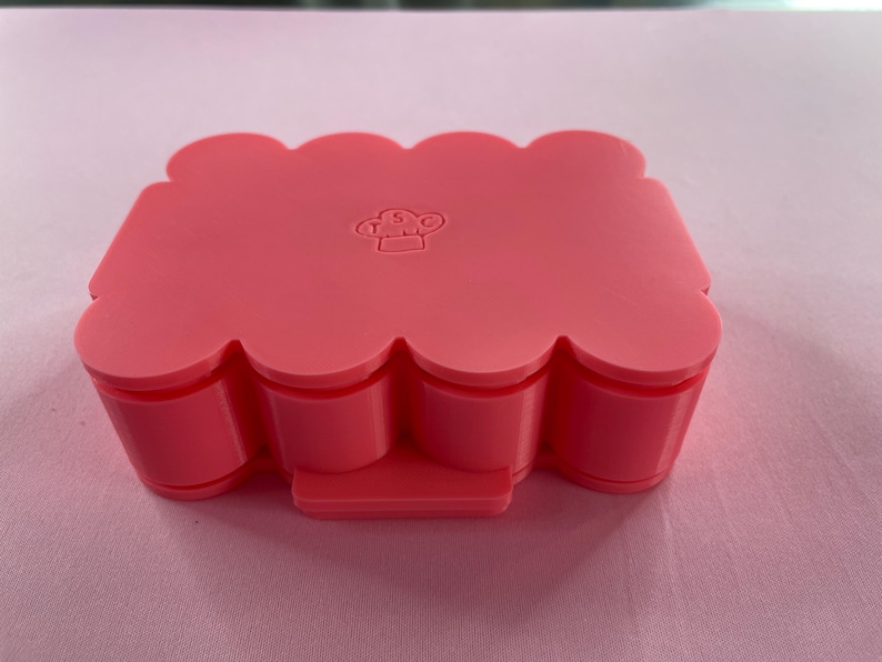May include: Pink silicone mold with a wavy edge and a small chef logo in the center. The mold is designed for making multiple small cakes or pastries.