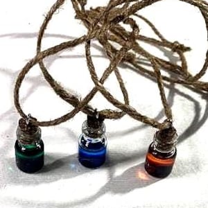 Op de afbeelding: Drie kleine glazen flesjes met kurken stoppen, elk gevuld met een andere kleur vloeistof: groen, blauw en oranje. De flesjes hangen aan een bruine touwketting, wat een uniek en kleurrijk accessoire creëert.