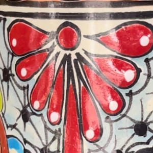 Könnte beinhalten: Nahaufnahme eines handbemalten Keramikvasenabschnitts. Das Design zeigt ein zentrales rotes Blumenmotiv mit weißen Akzenten, umgeben von einem hellblauen Hintergrund mit schwarzen spinnennetzartigen Mustern. Der obere Teil hat orange und schwarze geschwungene Formen.