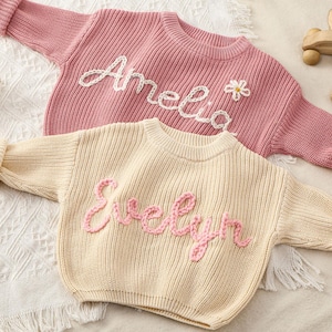 Könnte beinhalten: Zwei gestrickte Baby-Pullover, einer rosa und einer cremefarben, mit den Namen "Amelia" und "Evelyn" darauf gestickt. Der rosa Pullover hat ein kleines Blumendetail. Ein Häschen-Rassel und Holzspielzeug sind ebenfalls zu sehen.