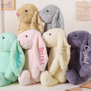 May include: A collection of plush bunny rabbits in various pastel colors, including mint green, pink, yellow, and purple. Each bunny has long, floppy ears with a name embroidered on one ear. The bunnies are soft and cuddly, perfect for children.