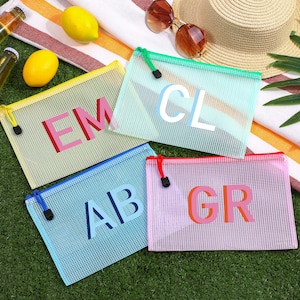 May include: Four rectangular mesh zipper pouches in yellow, light green, light blue, and pink. Each pouch features two large capital letters in contrasting colors: EM, CL, AB, and GR. The pouches have matching colored zipper pulls with black toggles.