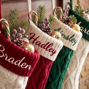May include: Four cable-knit Christmas stockings in red, white, and green, each featuring a personalized name embroidered on the cuff: Braden, Hadley, Ainsley, and Marley. Each stocking is filled with candy canes, pine sprigs, and ornaments.