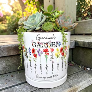 May include: A white ceramic planter filled with succulents and trailing plants. The planter is decorated with colorful floral illustrations and the text "Grandma's GARDEN" and "Growing since 1995". Names are written below the flowers.