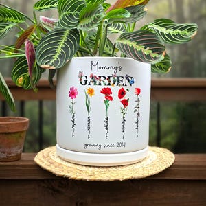 May include: A white ceramic planter with the text "Mommy's GARDEN" and colorful floral illustrations. The planter holds a green and red plant. The text "growing since 2014" is also visible.