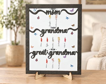 May include: A rectangular wooden frame containing a white board with black laser-cut text reading FIRST mom THEN grandma NOW great grandma. The board features colourful floral illustrations and butterfly graphics with names printed below each flower.
