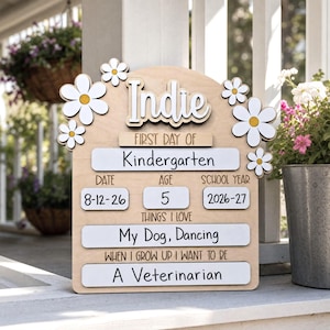 May include: A light-colored wooden sign with white daisy accents featuring the text Indie, First Day of Kindergarten, Date 8-12-26, Age 5, School Year 2026-27, Things I Love My Dog, Dancing, and When I Grow Up I Want To Be A Veterinarian.