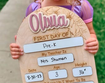 Puede incluir: Cartel de madera del primer día de escuela con el nombre "Olivia" en rosa. El cartel incluye campos para "Pre-K", "Mrs. Shuman", edad 3, altura 91.4 cm, fecha 8-30-23 y "A Doctor". Diseño floral en la parte superior.