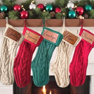 May include: Five personalized Christmas stockings in cream, red, and green, with cable knit design. Each stocking has a leather patch with a name: Daddy, Mommy, Kamdyn, Kotton, and Olivia. Festive holiday decor.