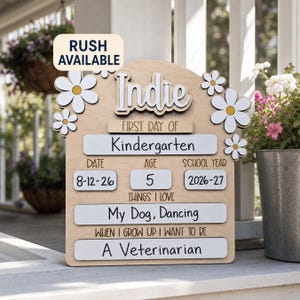 May include: A light-colored wooden sign with white daisy accents, featuring the text RUSH AVAILABLE, Indie, FIRST DAY OF, Kindergarten, DATE 8-12-26, AGE 5, SCHOOL YEAR 2026-27, THINGS I LOVE My Dog, Dancing, and WHEN I GROW UP I WANT TO BE A Veterinarian.