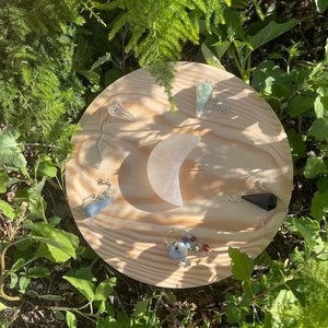 May include: A wooden board with a crescent moon shape and several crystal pendulums with chains. The pendulums are made of various crystals, including clear quartz, amethyst, and black obsidian.