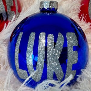 May include: Three spherical Christmas ornaments with silver glitter lettering. Two red ornaments feature the names MICHAEL and OZZIE, while the central blue ornament displays the name LUKE. Each ornament has a silver metal cap and loop.