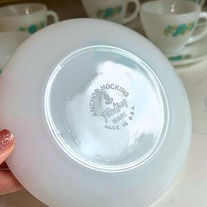 May include: White saucer with the Anchor Hocking Fire King Ware logo. The saucer is part of a vintage tea set, with other cups and saucers visible in the background. The tea set has a floral design.