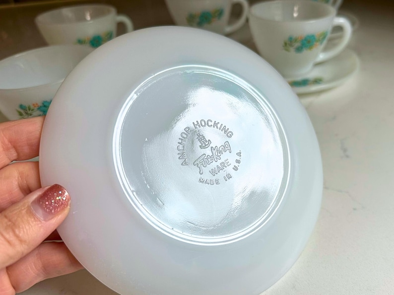 May include: White saucer with the Anchor Hocking Fire King Ware logo. The saucer is part of a vintage tea set, with other cups and saucers visible in the background. The tea set has a floral design.