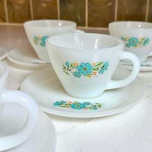 May include: White teacups and saucers with a floral design in turquoise and orange. The cups have a rounded shape with a handle, and the saucers are round. The teacups and saucers are arranged on a white surface.