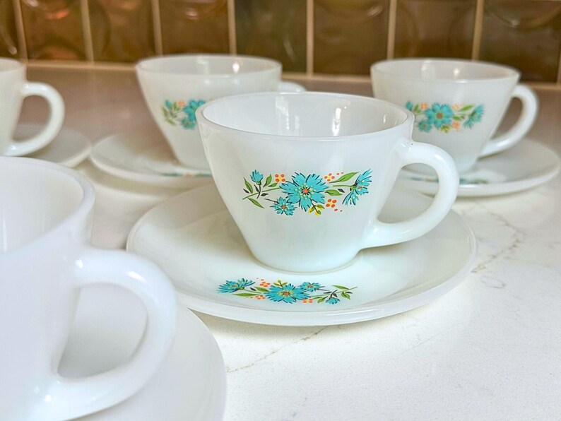May include: White teacups and saucers with a floral design in turquoise and orange. The cups have a rounded shape with a handle, and the saucers are round. The teacups and saucers are arranged on a white surface.