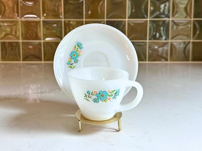 May include: White teacup and saucer set with a floral design in turquoise and orange. The cup and saucer are displayed on a small gold-colored stand. The set is likely vintage or retro, suitable for serving tea or coffee.