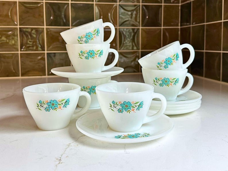 May include: A set of white teacups and saucers with a floral design in turquoise and orange. The cups have a rounded shape with handles, and the saucers are round. The set is arranged on a white surface.