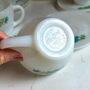 May include: White teacup and saucer set with a floral design in turquoise and green. The teacup has a curved handle and the saucer is round. The bottom of the teacup is stamped with "Oven Fire King Ware USA Made".