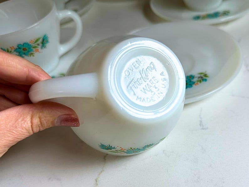 May include: White teacup and saucer set with a floral design in turquoise and green. The teacup has a curved handle and the saucer is round. The bottom of the teacup is stamped with "Oven Fire King Ware USA Made".