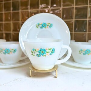 May include: A set of white teacups and saucers with a floral design in turquoise and orange. The cups and saucers are arranged on a white surface, with a gold-colored stand holding one cup. The background is a brown tile wall.