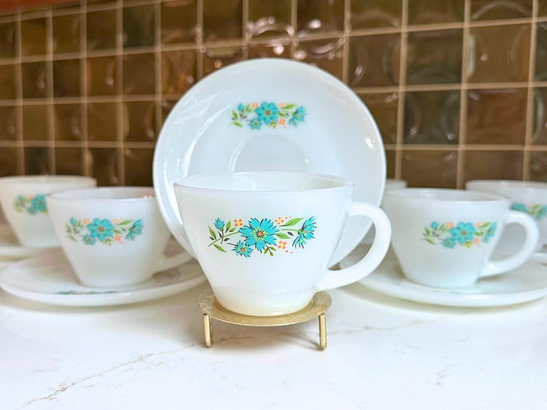 May include: A set of white teacups and saucers with a floral design in turquoise and orange. The cups and saucers are arranged on a white surface, with a gold-colored stand holding one cup. The background is a brown tile wall.