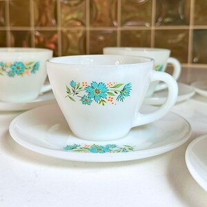 May include: White teacups and saucers with a floral design in turquoise and orange. The cups have a rounded shape with a handle, and the saucers are round. The floral pattern is repeated on both the cups and saucers.