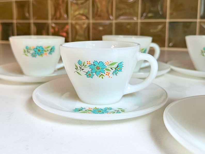 May include: White teacups and saucers with a floral design in turquoise and orange. The cups have a rounded shape with a handle, and the saucers are round. The floral pattern is repeated on both the cups and saucers.