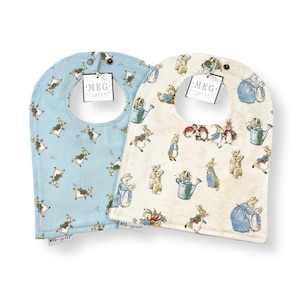 May include: Two baby bibs with a Peter Rabbit design. One bib is blue with white rabbits and the other is cream with brown rabbits. Both bibs have a white tag with the text "MEG - original".