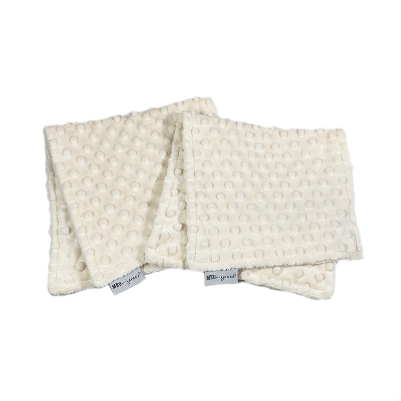 May include: Four ivory-colored, square baby washcloths with a textured, dotted surface. Each cloth has a small, rectangular tag with the text "MEG...yup" printed on it. The washcloths are stacked and folded.