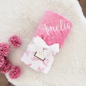 May include: A pink baby blanket with the name "Amelia" embroidered in white script. The blanket is tied with a white ribbon and a tag that reads "MEG original". The blanket has a pink bow and polka dot pattern.