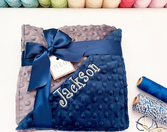 May include: A gray and navy blue baby blanket with a navy blue satin ribbon tied in a bow. The blanket is personalized with the name "Jackson" in white embroidery. The blanket is wrapped and ready for gifting.