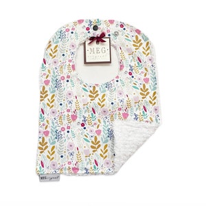 May include: A white bib with a floral pattern in pink, yellow, and blue. The bib has a white terry cloth backing and a snap closure. The bib has a label that says "MEG original".