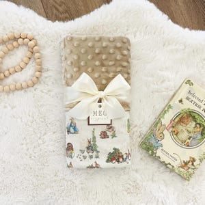 May include: A beige baby blanket with a cream-colored bow and a tag that reads "MEG original." The blanket is paired with a vintage copy of "Selected Beatrix Potter." The blanket features a patterned fabric with illustrations of rabbits and other woodland creatures.