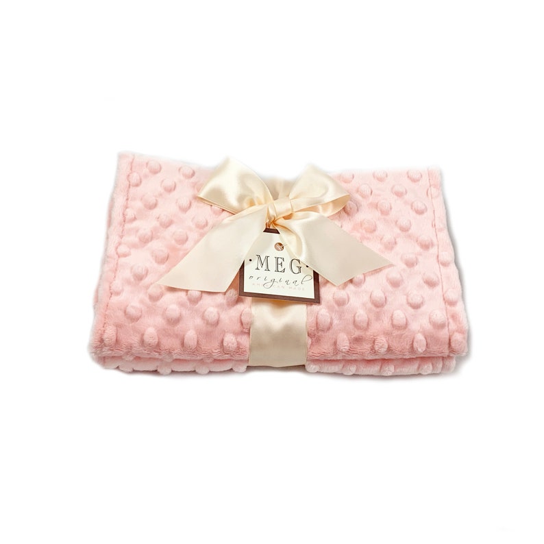 May include: A soft, pink baby blanket with a textured dot pattern. The blanket is tied with a cream-colored satin ribbon and bow. A tag with the text "MEG original" is attached to the ribbon.