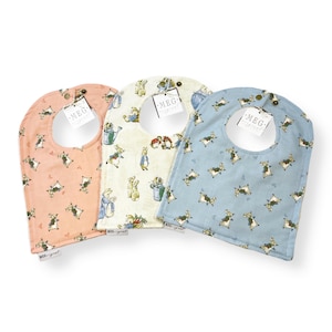 May include: Three baby bibs with Peter Rabbit designs. The bibs are in pink, white, and blue with a Peter Rabbit pattern. Each bib has a snap closure.