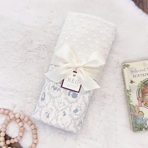 Może przedstawiać: Biały kocyk dla dziecka z teksturowaną, kropkowaną powierzchnią jest zawiązany kremową satynową wstążką. Dolna część ma niebiesko-biały wzór z ilustracjami królików i innych leśnych stworzeń. Etykieta głosi "MEG original".