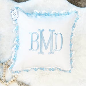 May include: White square cushion with light blue embroidered monogram BMD. The cushion has a light blue bow-shaped trim around the edges. A tag in the upper left corner reads "MEG ORIGINAL".
