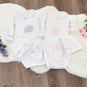 Puede incluir: Dos vestidos de bebé blancos con gorros y calcetines a juego. Un vestido tiene un monograma azul, el otro un monograma rosa. Cada vestido tiene un cuello y puños con volantes. Los gorros y calcetines están adornados con encaje y lazos.