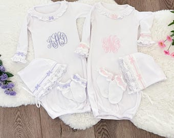 May include: Two white baby gowns with matching bonnets and socks. One gown has a blue monogram, the other a pink monogram. Each gown has a ruffled collar and cuffs. The bonnets and socks are trimmed with lace and bows.