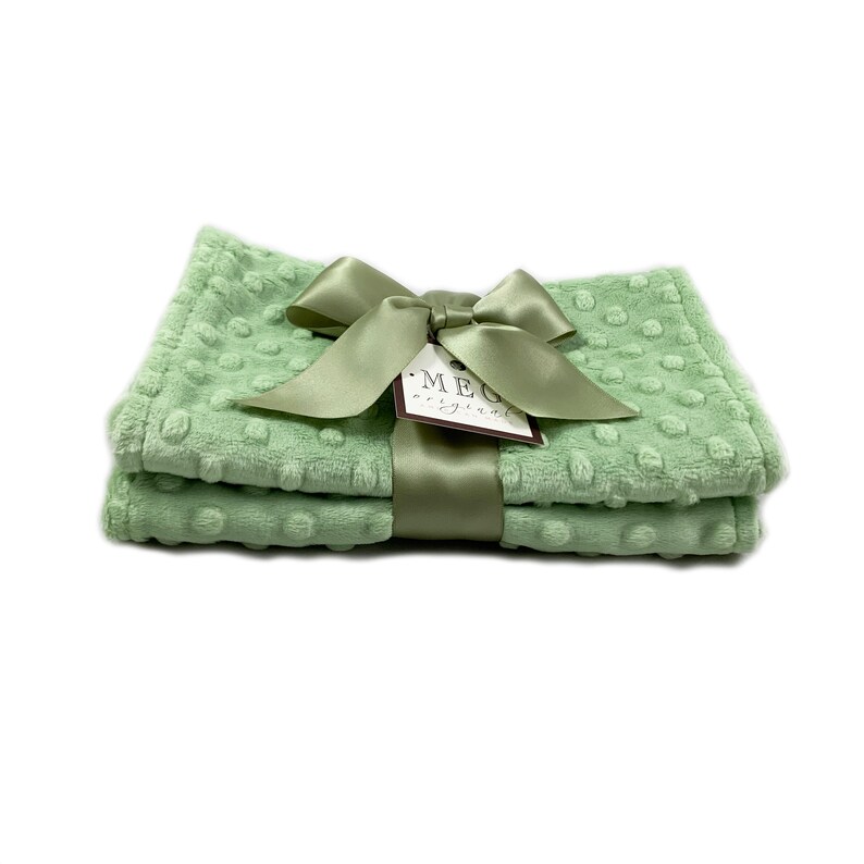 May include: Two sage green baby blankets with a raised dot pattern, tied with a matching satin ribbon and bow. A tag with the text "MEG original" is attached to the ribbon. The blankets are folded and stacked.
