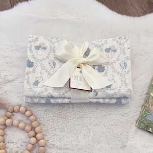 May include: A folded baby blanket with a light blue and white pattern featuring rabbits and floral designs. The blanket is tied with a cream-colored ribbon and a tag that reads "ME original".