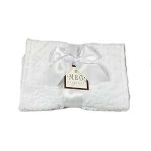 May include: A white baby blanket with a textured dot pattern, tied with a white satin ribbon and bow. A brown tag with the text "MEG original hand made" is attached to the ribbon.